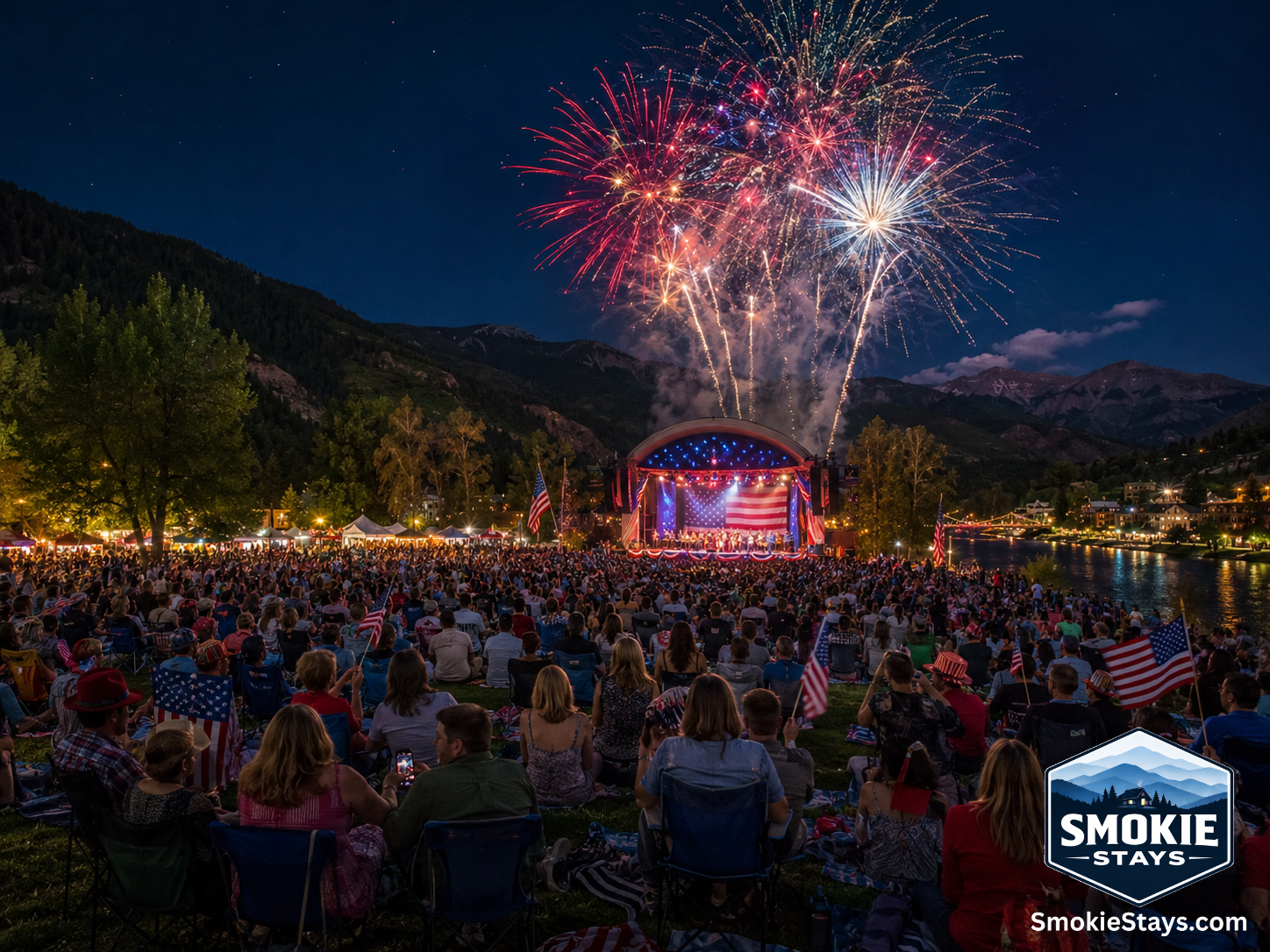 A large crowd gathers on the lawn at an outdoor Fourth of July concert in Pigeon Forge, Tennessee, with fireworks bursting above the stage and the Smoky Mountains in the background.